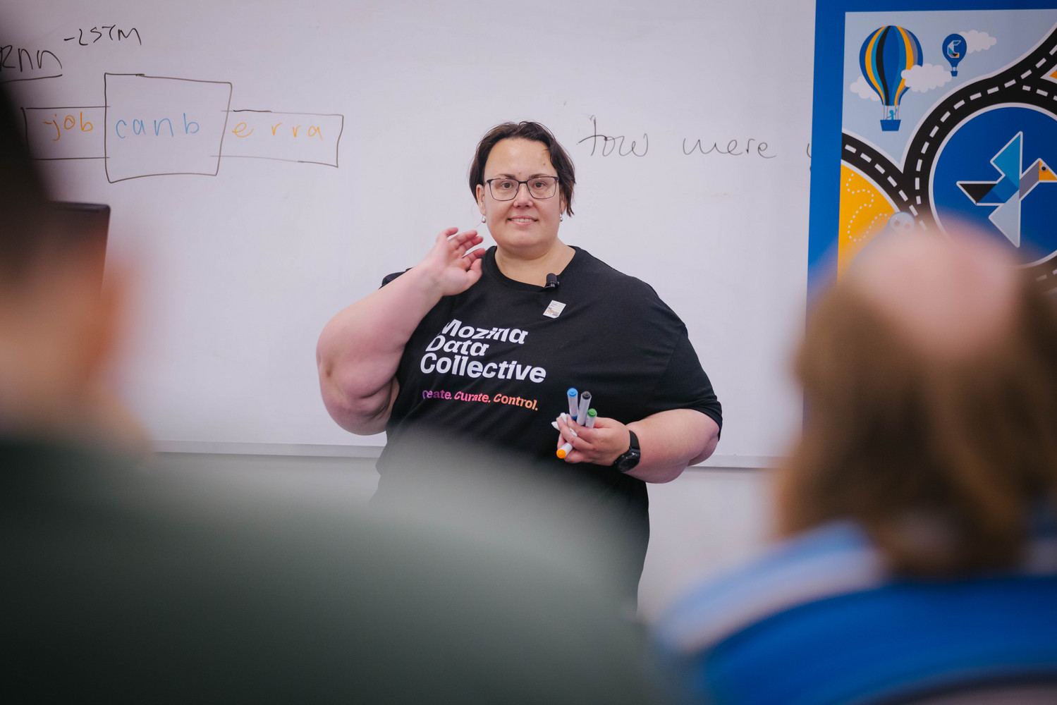 Kathy Reid, wearing a Mozilla Data Collective t-shirt and holding whiteboard markers, speaks to a room of tutorial participants in front of a whiteboard, with a colourful conference banner to one side.